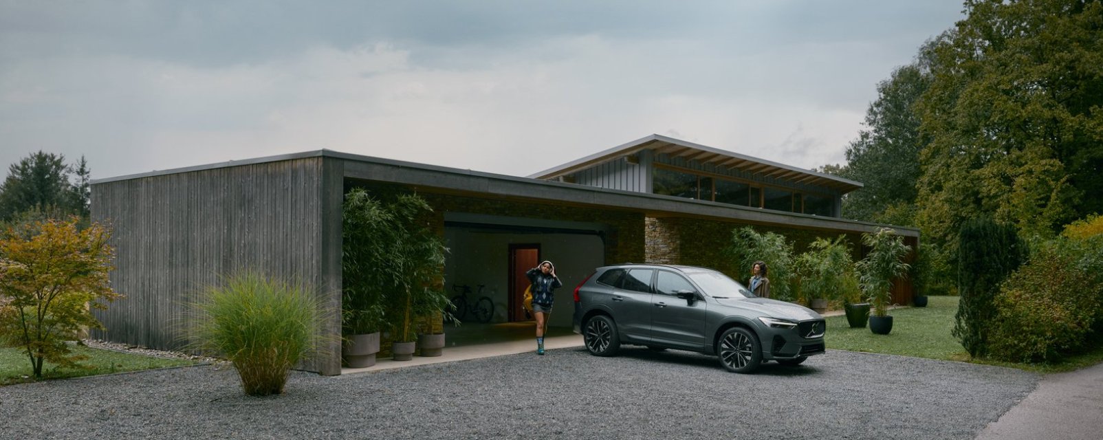 SUV hibrida Volvo XC60 color gris estacionada en el porche de una casa con una mujer caminando al lado.